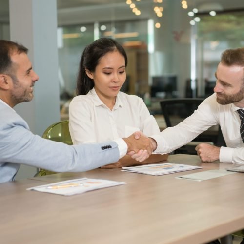 Business people shaking hands at desk in office. Multiethnic businesspeople meeting and sitting with modern blurred interior in background. Agreement concept.